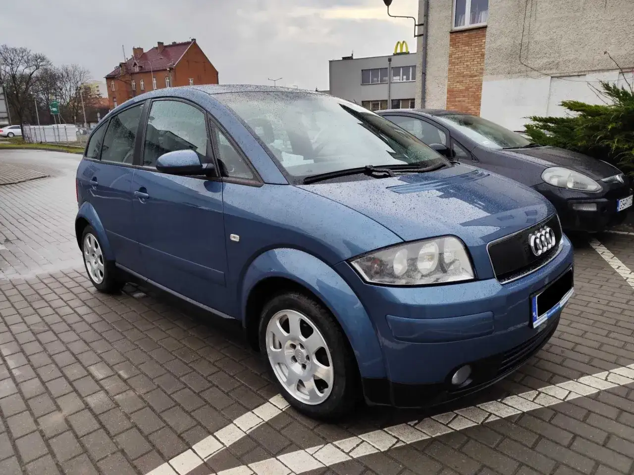 Niebieski Audi A2 1.4 benzyna na mokrym parkingu.