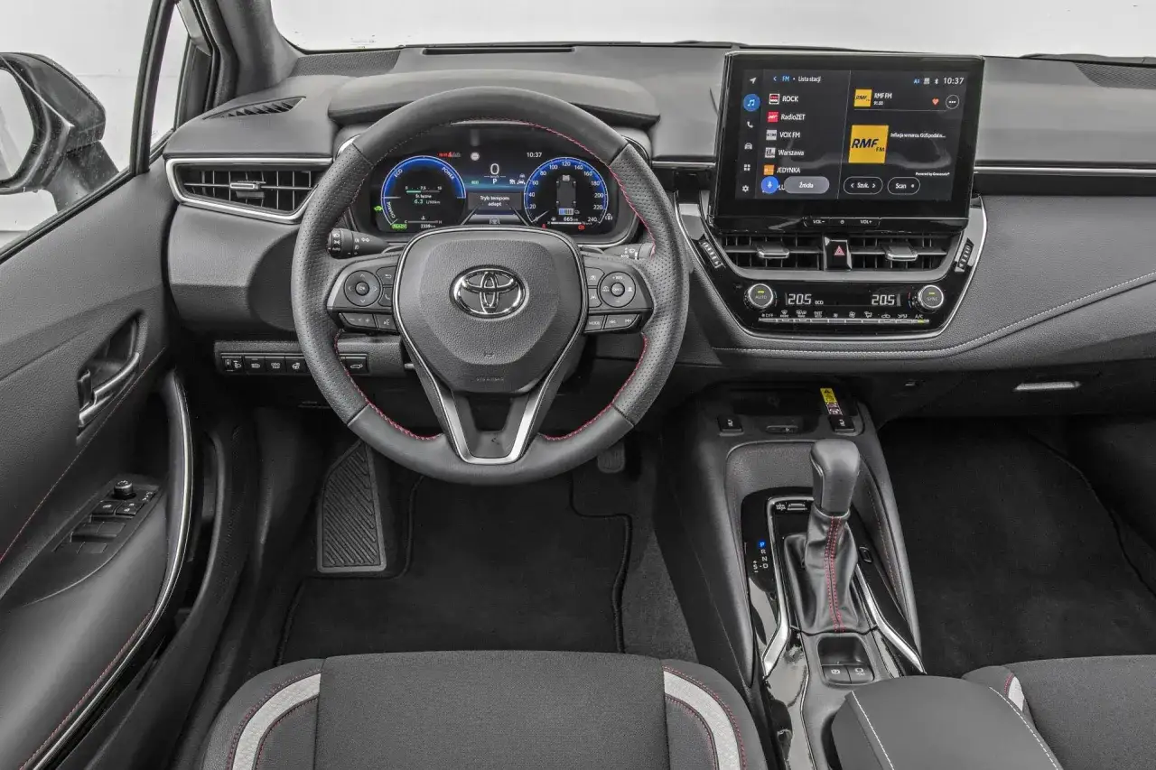 Wnętrze Toyoty Corolla sedan: kierownica, ekran multimediów z radiem RMF FM i nawigacją, cyfrowe zegary.