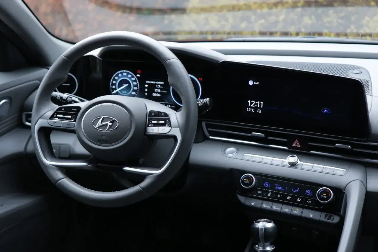 Wnętrze Hyundaia Elantry z cyfrowymi zegarami i ekranem multimediów. Kierownica z przyciskami, widoczna dźwignia zmiany biegów. Wiele osób szuka hyundai elantra opinie przed zakupem.