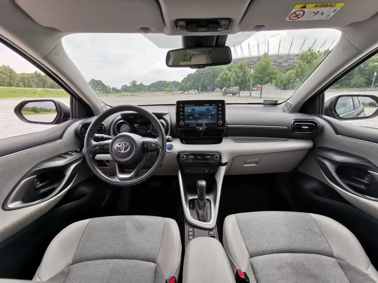 Wnętrze Toyoty Yaris 4 z widokiem na stadion. Jasna tapicerka, ekran multimedialny i kierownica z logo producenta.