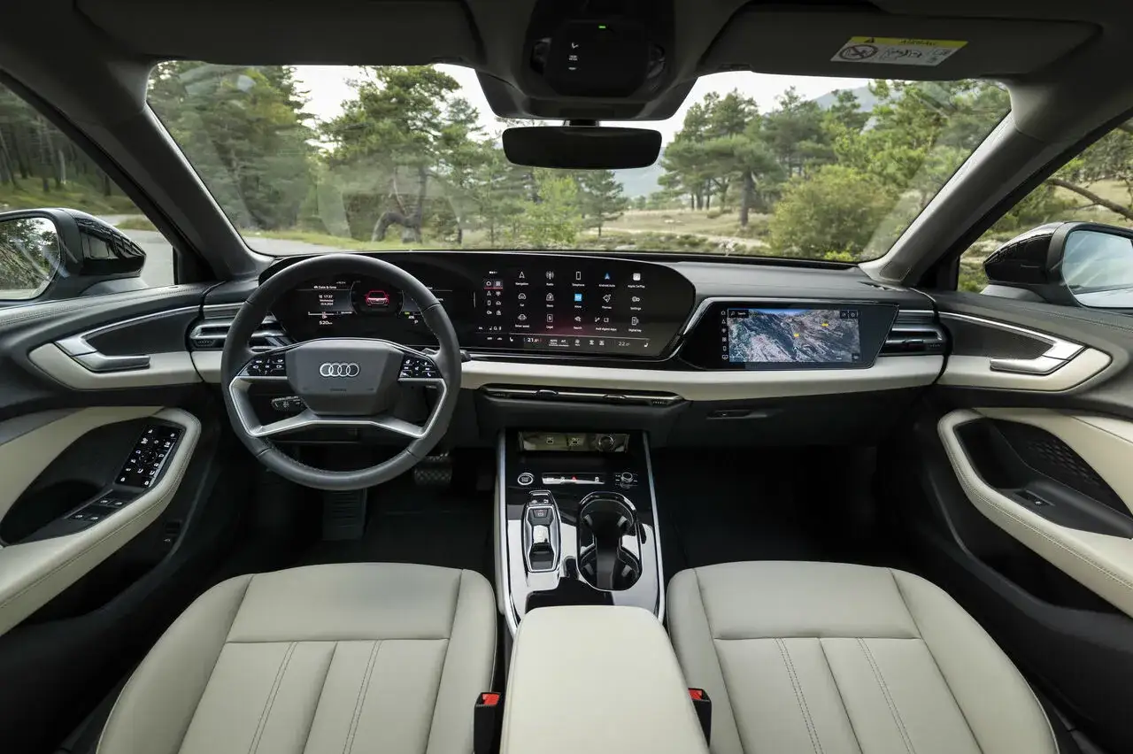 Wnętrze Audi A5 B10 z nowoczesnym kokpitem, dużymi ekranami i jasnymi skórzanymi fotelami. Widok z perspektywy kierowcy na drogę wśród drzew.