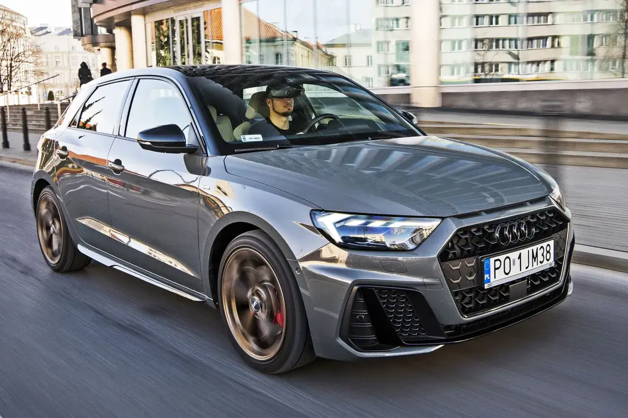 Szary Audi A1 Sportback w ruchu miejskim. Kierowca w goglach VR. Kompaktowe a1 wymiary idealne do miasta.