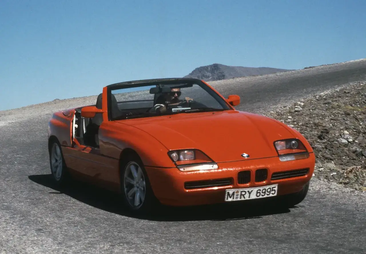 Pomarańczowy kabriolet BMW Z1 z opuszczonymi drzwiami jedzie po krętej drodze w górach.