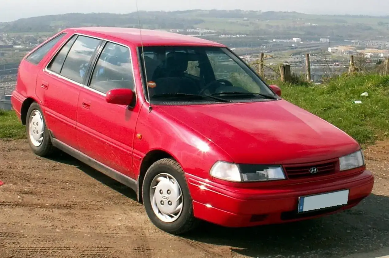 Czerwony Hyundai Pony z lat 90. stoi na błotnistej drodze, z widokiem na dolinę i miasto w tle.