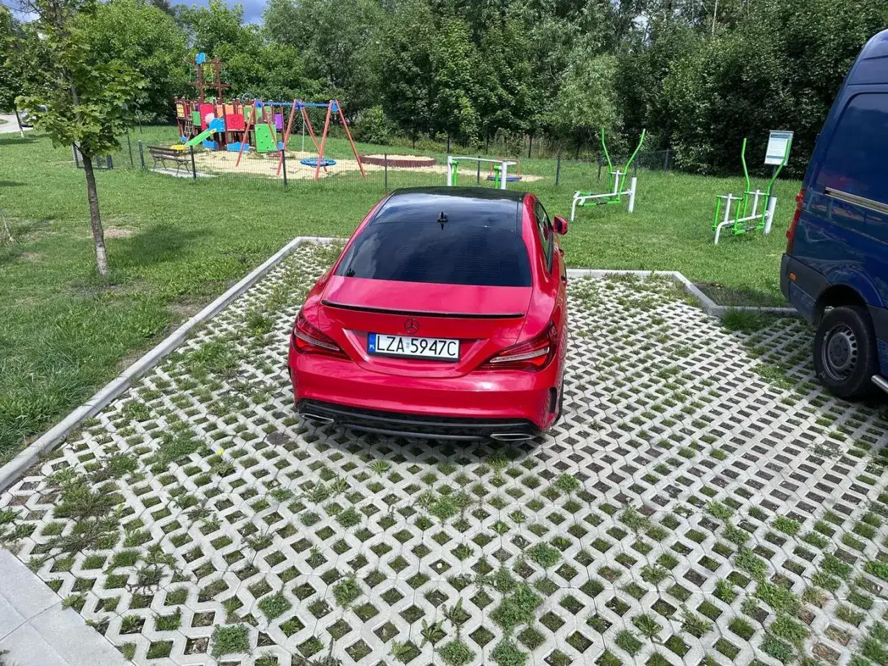 Czerwony Mercedes z tablicą LZA 5947C zaparkowany na ekologicznym parkingu obok placu zabaw i siłowni plenerowej.