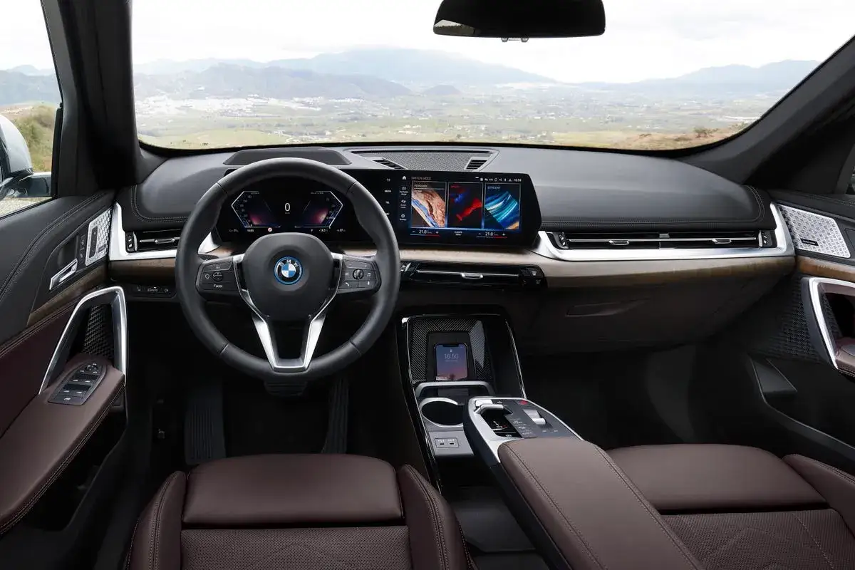 Wnętrze BMW X1 elektryk: skórzana tapicerka, nowoczesny kokpit z dwoma ekranami i kierownicą. Widok na górzysty krajobraz.