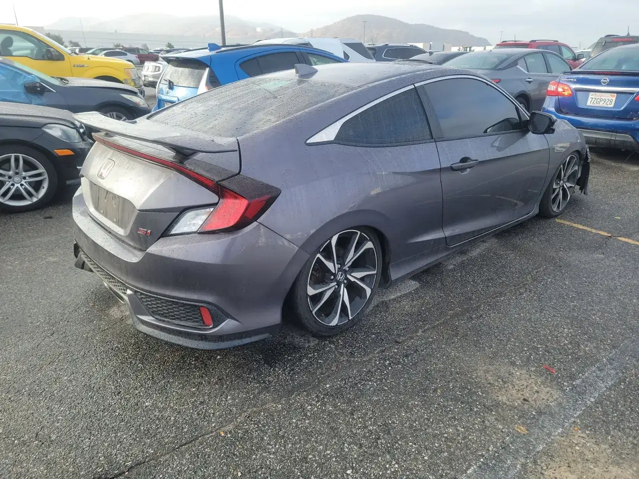 Szary Honda Civic Si coupe z czarnym spojlerem i felgami stoi na mokrym parkingu.