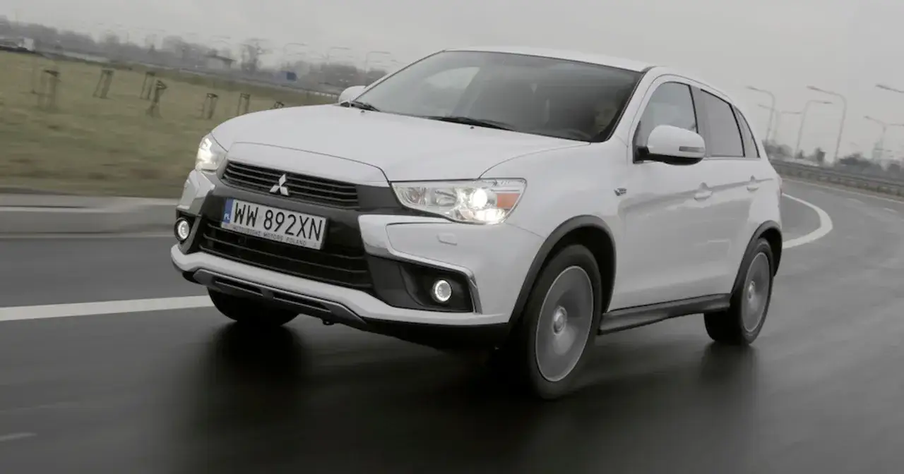 Białe Mitsubishi ASX 1.6 benzyna na mokrej drodze, z widocznymi światłami i tablicą rejestracyjną WW 892XN.