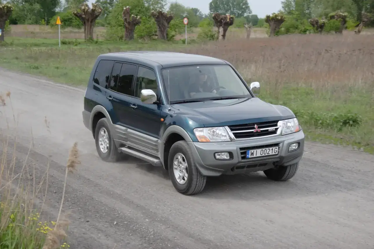 Zielony Mitsubishi Pajero 3 jedzie po szutrowej drodze, wzbijając kurz. W tle widać drzewa i pola.