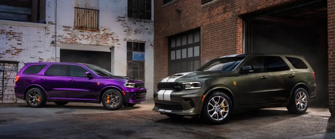 Dwa Dodge Durango, jeden fioletowy, drugi zielony, stoją przed ceglanymi budynkami. Poznaj imponujące dodge durango wymiary i możliwości tych SUV-ów.