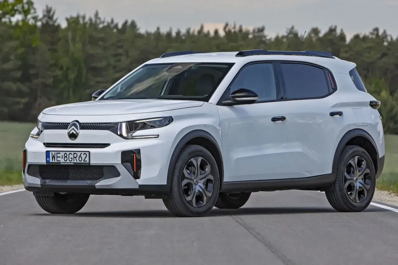 Biały, mały SUV Citroën C3 Aircross z czarnymi relingami dachowymi i przyciemnianymi szybami jedzie po asfaltowej drodze.