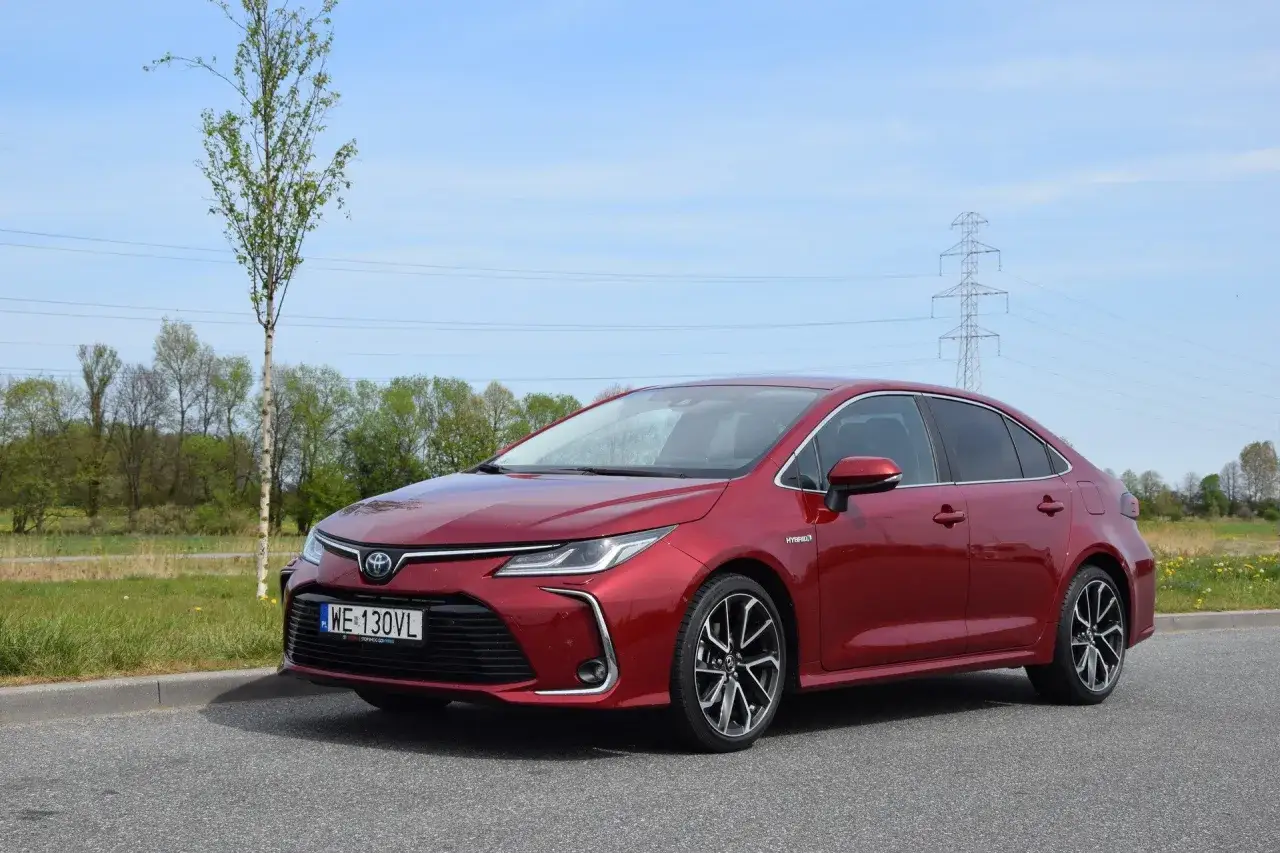 Czerwona Toyota Corolla 1.8 Hybrid zaparkowana na poboczu drogi, z drzewem i liniami energetycznymi w tle.