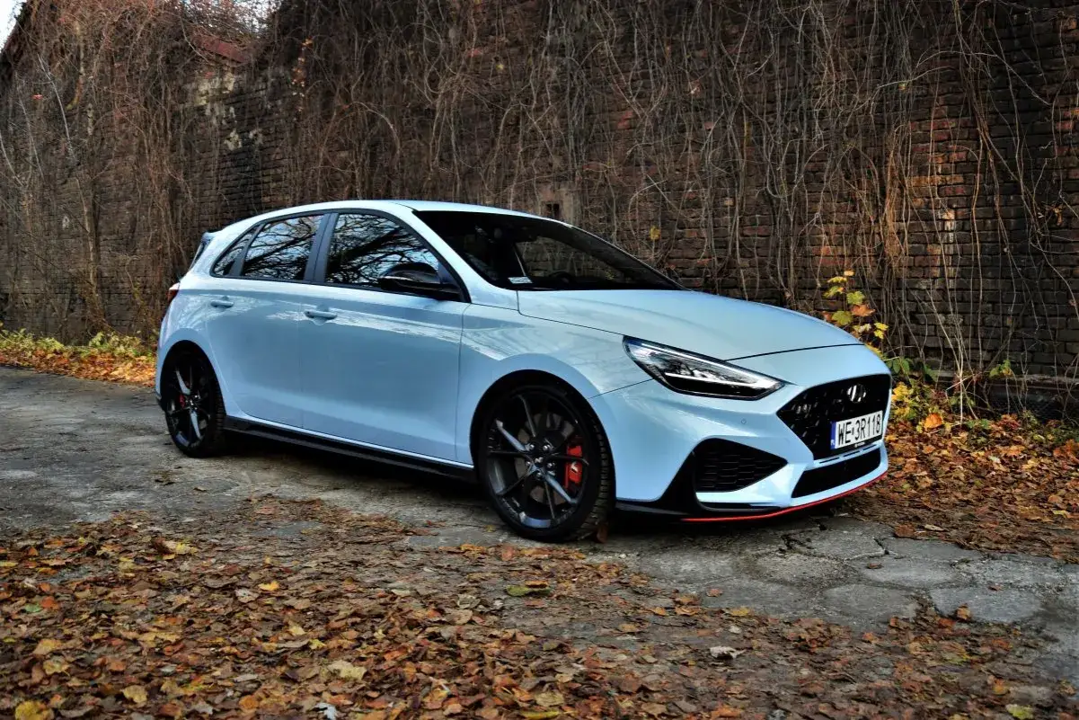 Jasny Hyundai i30 N z czarnymi felgami i czerwonymi zaciskami hamulcowymi stoi na tle ceglanej ściany porośniętej dzikim winem.