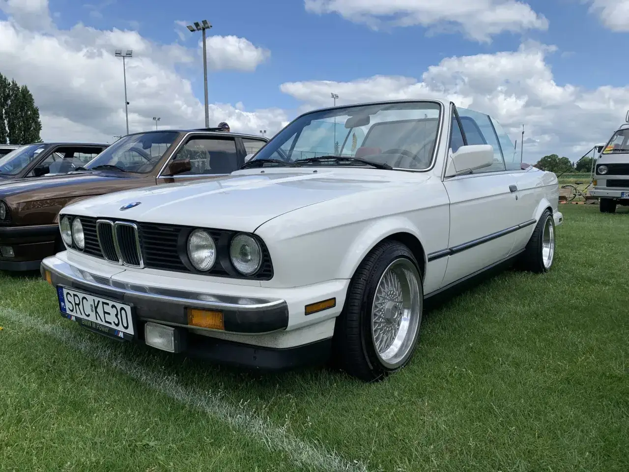 Białe BMW 325i kabriolet z chromowanymi felgami stoi na trawie pod błękitnym niebem z chmurami.