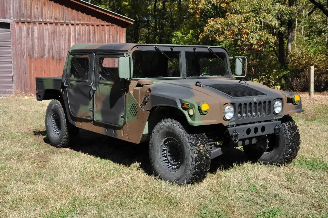 Potężny Hummer H1 w kamuflażu, gotowy na każdą terenową przygodę, stoi na trawie obok drewnianej szopy.