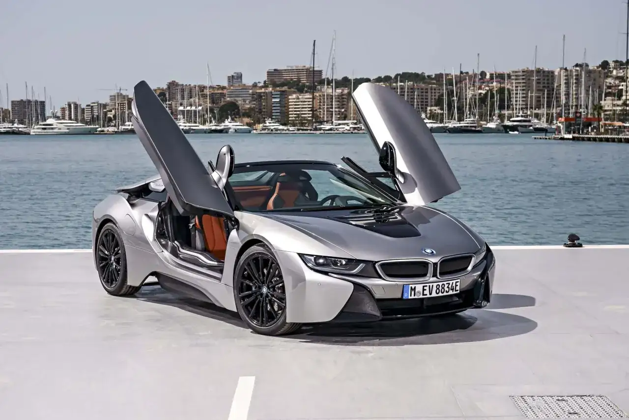 Srebrne BMW i8 z otwartymi drzwiami, stojące przy nabrzeżu z jachtami w tle.