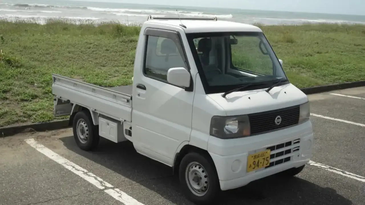 Biały kei truck z otwartą paką stoi na parkingu przy plaży.