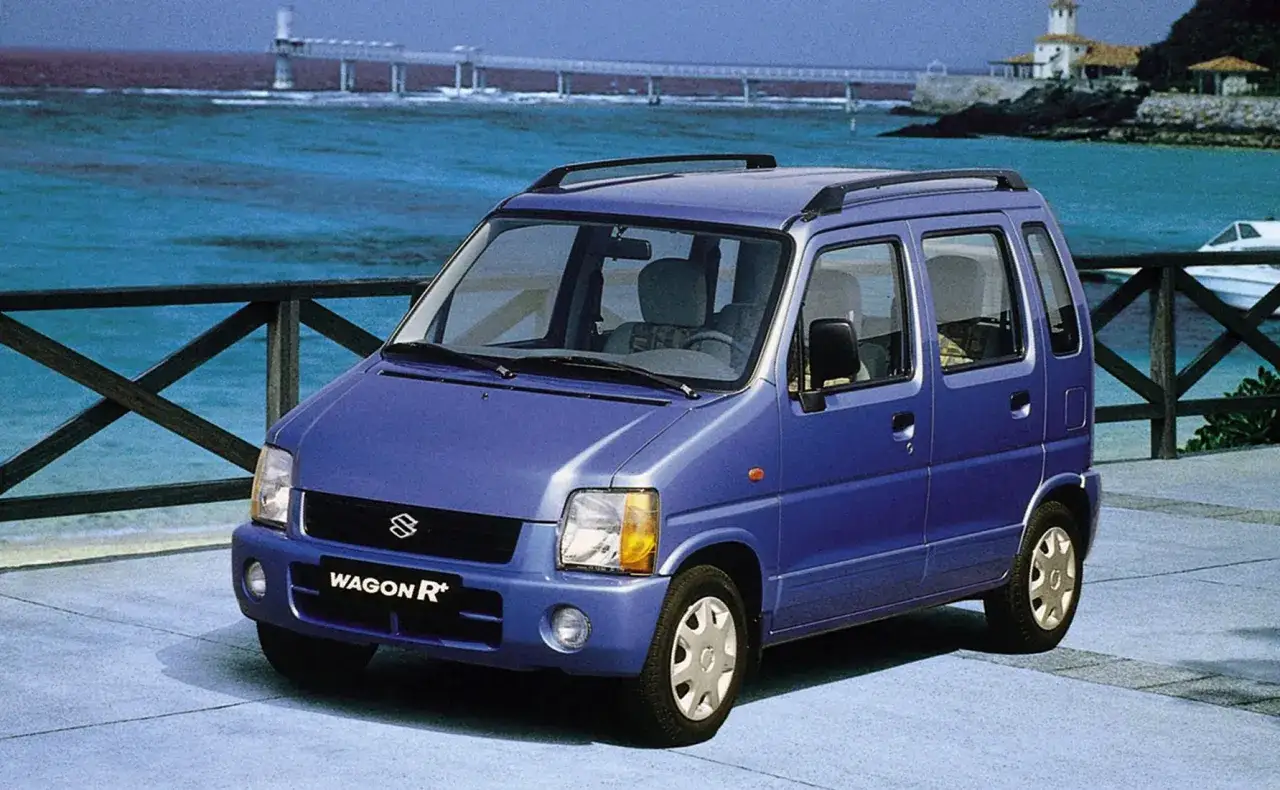 Niebieski Suzuki Wagon R+ zaparkowany nad morzem, z widokiem na most i budynki w oddali.