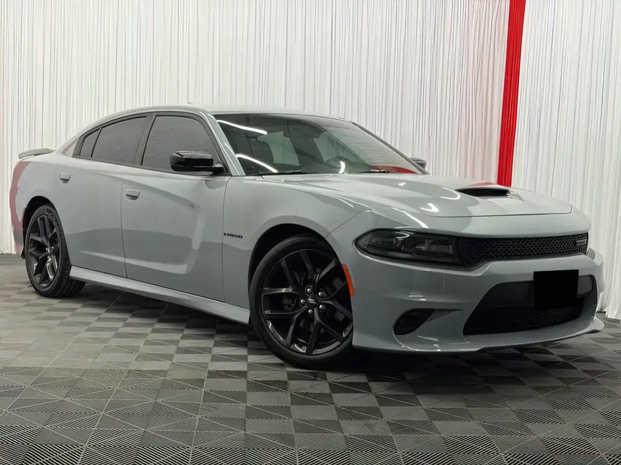 Szary Dodge Charger, marka samochodu na d, z czarnymi felgami i przyciemnianymi szybami, stoi na kratkowanej podłodze.