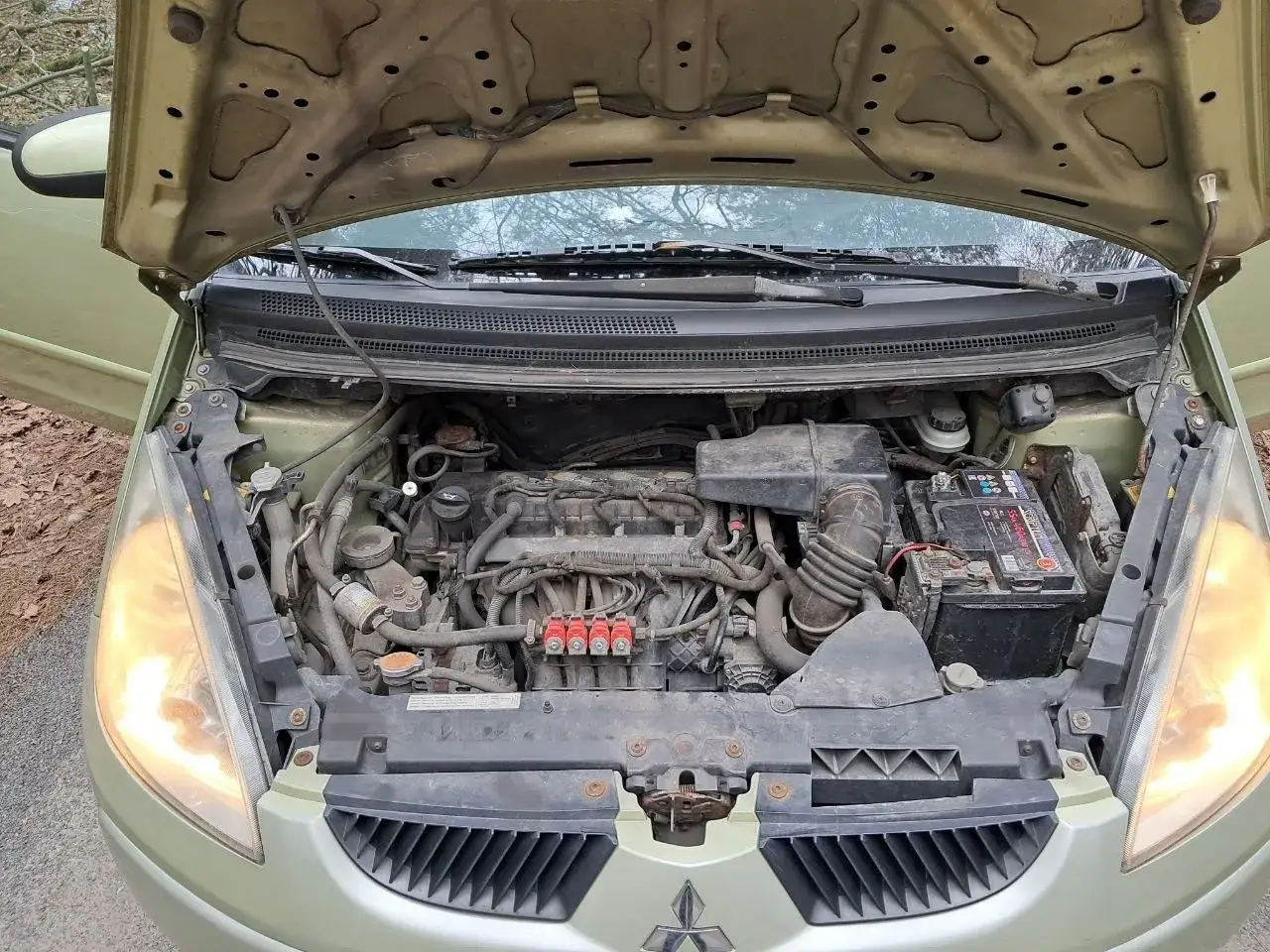 Silnik Mitsubishi Colt 1.3 z otwartą maską. Widoczny akumulator, przewody i elementy układu paliwowego.