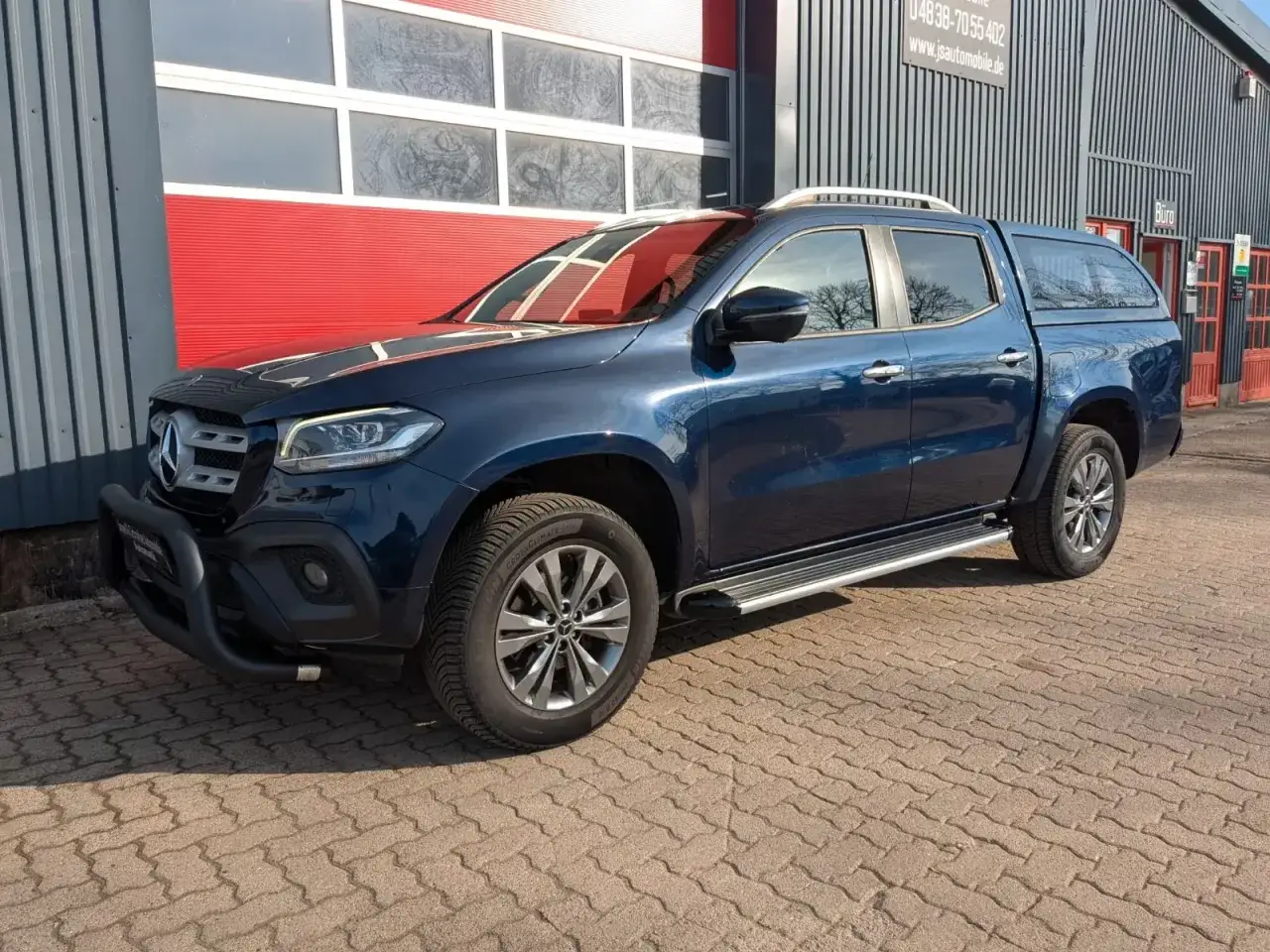 Ciemnoniebieski mercedes pickup z zabudową na pace, zaparkowany na kostce brukowej przed budynkiem z czerwonymi drzwiami.