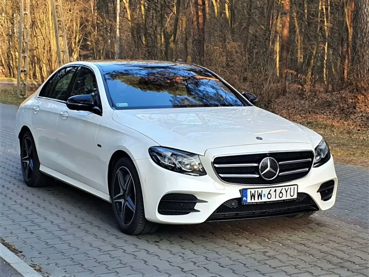 Biały Mercedes E300 z czarnym dachem i lusterkami, zaparkowany na brukowanej ścieżce w lesie.