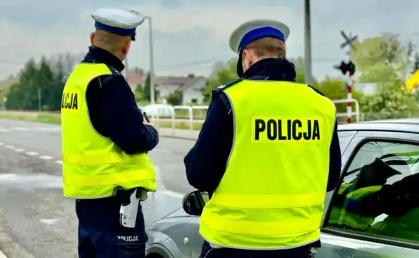 Dwóch policjantów w kamizelkach odblaskowych przy samochodzie. Jeden z nich trzyma broń. Prawdopodobnie zatrzymali kierowcę za przekroczenie prędkości o 50 km/h.