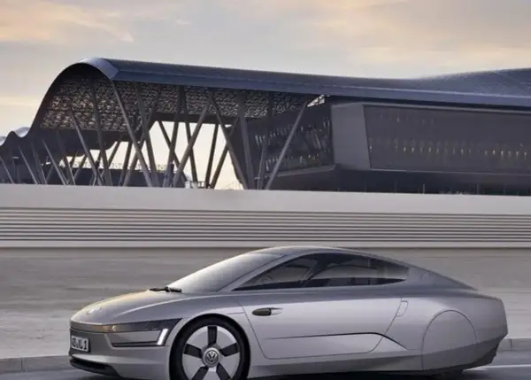 Srebrny, futurystyczny vw xl1 z charakterystycznym dachem i nowoczesnym budynkiem w tle.