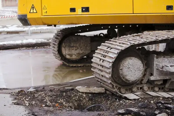 Gąsienice koparki, z widoczną zwolnicą, stoją na błotnistym terenie po pracach drogowych.