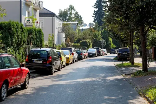 Ulica z wieloma samochodami, gdzie parkowanie na chodniku jest powszechne. Widać domy i drzewa.