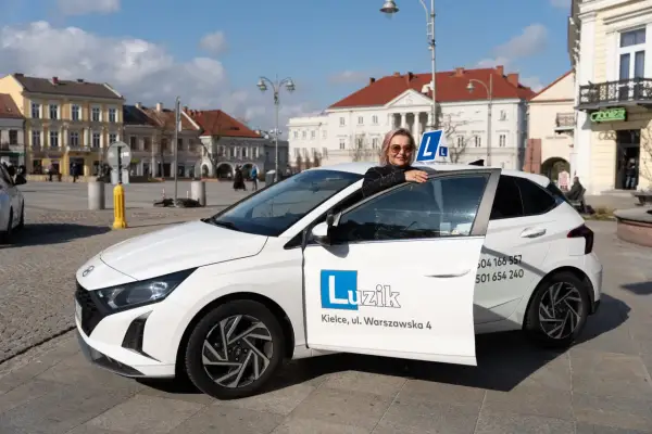 Kobieta z uśmiechem opiera się o drzwi białego samochodu szkoleniowego z literą "L" na dachu. To idealny moment, by zacząć naukę jazdy i zdobyć prawo jazdy B.