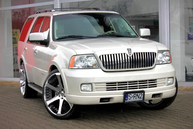Biały Lincoln Navigator 2024 z chromowanymi felgami, zaparkowany na mokrym bruku przed budynkiem z dużymi oknami.